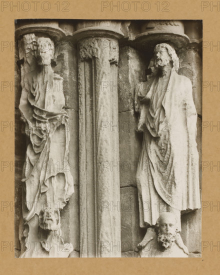 Fürstentor, Ap. r.3.4.v.NO., Detail of the Princely Portal in Bamberg Cathedral, album page, photograph, This photo is part of an album., number: '85', Bamberg, 1900 - c. 1920, photographer, Bavaria, paper, baryta paper, gelatin silver print, developed gelatin silver print, height 208 mm x width 164 mm, church (exterior), Bamberg Cathedral, A close-up view of a stone relief framed by a rectangular border, showing two vertical fluted columns in the center with carved capitals, flanked by carved human figures in draped garments; on the left a weathered figure with a long draped robe and an eroded face, and below it a smaller carved head with a beard, while on the right a standing robed figure with a bearded head and detailed folds in the fabric, and below that another carved bearded head with a distinct floral-like beard detail; the stone surface shows texture, wear, and subtle shadows emphasizing the carved folds and architectural elements.