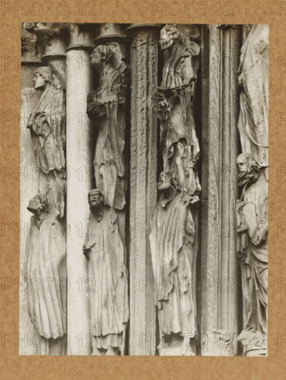 Detail of the Princely Portal in Bamberg Cathedral, Princely Gate Statue no. 1.2.3. vN, album page, photograph, This photo is part of an album., number: '91', Bamberg, 1900 - c. 1920, photographer, Bavaria, paper, baryta paper, gelatin silver print, gelatin silver developing print, height 211 mm x width 158 mm, church (exterior), Bamberg Cathedral, A sepia-toned photograph of a carved stone façade showing a vertical arrangement of elongated human figures in robes positioned between columns and narrow pilasters; the figures display varied head poses and facial features with some heads turned in profile and others facing forward, and the drapery of their garments forms deep vertical folds and textured surfaces; the stone shows weathering and roughness with cracks and chips visible along edges, and narrow shadowed recesses separate the figures and architectural elements; a textured paper border frames the image.