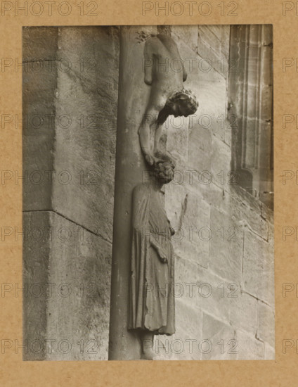 Detail of the Princely Portal in Bamberg Cathedral, Princely Gate Judas Column, the figure, album page, photograph, This photo is part of an album., number: '94.', Bamberg, 1900 - c. 1920, photographer, Bavaria, paper, baryta paper, gelatin silver print, height 226 mm x width 162 mm, church (exterior), Bamberg Cathedral, A grayscale photograph shows a carved stone figure attached to a vertical column against a textured stone wall; the figure is draped in a long garment with visible folds and stands upright while another smaller carved figure appears above, bending forward with one arm extended downward toward the head of the lower figure; the upper figure has stylized hair and a rounded form, and the lower figure's facial features and drapery are visible; the stone surfaces show weathering, grain, and subtle discoloration, and the image is bordered by a narrow lighter frame and a wider textured mat.
