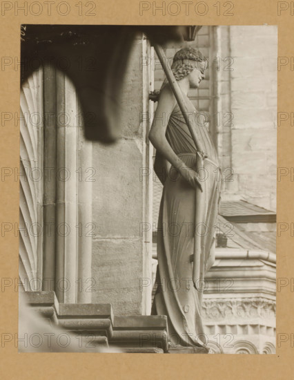 Fürstentor, Synagogue page, Detail of the Princely Portal in Bamberg Cathedral, album page, photograph, This photo is part of an album., number: '102', Bamberg, 1900 - c. 1920, photographer, Bavaria, paper, baryta paper, gelatin silver print, developed gelatin silver print, height 205 mm x width 161 mm, church (exterior), Bamberg Cathedral, A sepia-toned photograph showing a stone statue of a draped female figure in profile, positioned against a vertical stone pillar and holding a long diagonal staff that rests against her shoulder, with textured hair styled in waves and a blindfold across her eyes; the statue's flowing garment has visible folds and reaches down to a pedestal, while architectural details of the surrounding building appear in the background, including carved stone columns, layered molding, and a patterned facade partly visible through a window frame.