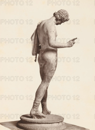 Museo di Napoli Narciso, Sculpture of Narcissus, album page, photograph, This photo is part of an album., workshop stamp: 'Giorgio Sommer, Largo Vittoria, Napoli', number: '1527', photographer: Giorgio Sommer, Naples, 1874 - in or before 1879, paper, albumen print, height 249 mm x width 184 mm, sculpture, (story of) Narcissus, A monochrome photograph of a classical marble statue shown in profile from behind, depicting a nude male figure wearing a wreath in his curled hair and a draped cloth hanging from one shoulder, with one arm extended forward and a pointing hand, smooth musculature and worn surface tones, detailed sandals with ornate patterns on the feet, and the figure standing on a round textured plinth against a plain light background.