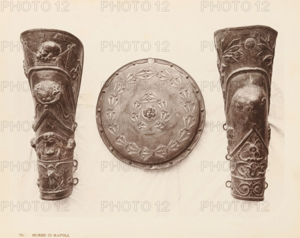 Decorated objects, Museo di Napoli Saffo, album page, photograph, This photo is part of an album., workshop stamp: 'Giorgio Sommer, Largo Vittoria, Napoli', number: '7517', photographer: Giorgio Sommer, Naples, 1874 - in or before 1879, paper, albumen print, height 202 mm x width 254 mm, military clothing and other equipment (uniforms, cap, armor, helmet, etc.), A centered round shield with raised decorative motifs and a small sculpted face at its center hangs between two tall, elaborately carved cylindrical objects; the shield shows a wreath-like pattern and small round studs along its rim. The left cylindrical object features multiple relief scenes including a bearded head near its top, ornate bands and panels with animal and human figures, and loops protruding from its side. The right cylindrical object mirrors the left in shape and also displays relief carvings including vegetal motifs near the top, a rounded protuberance with facial features set into a recessed panel, intertwined ...