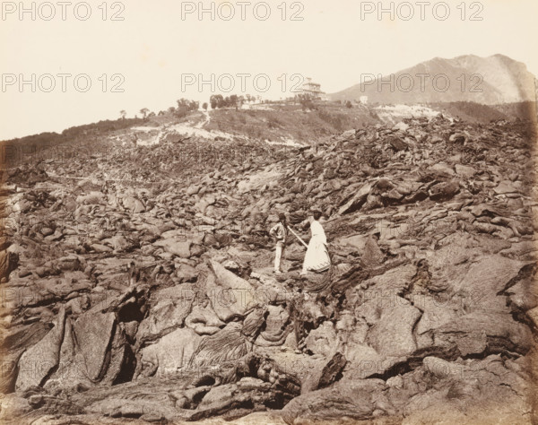 Lava of Vesuvius (Naples), Man and woman on the hardened lava after an eruption of Mount Vesuvius, album page, photograph, This photo is part of an album., number: '6139', workshop stamp: 'Giorgio Sommer, Largo Vittoria, Naples', photographer: Giorgio Sommer, Naples, 1874 - in or before 1879, paper, albumen print, height 203 mm x width 249 mm, historical persons, adult woman, adult man, cold lava, Mount Vesuvius, A wide rocky landscape fills the foreground with textured, folded formations and rough surfaces in muted tones, leading up to a ridge with sparse trees and a building complex perched near the top; two people stand among the rocks near the center, one wearing a light dress with a patterned hem and the other in lighter trousers and a jacket while holding a long pole, and a large mountain rises in the background to the right under a pale sky.