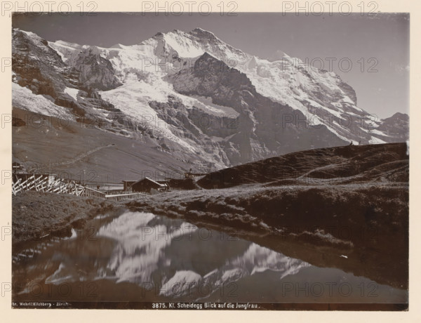 Jungfrau, seen from the Kleine Scheidegg, Kl. Scheidegg View of the Jungfrau, photograph, This photo is part of a cover., inscription: 'Gebr. Wehrli Kilchberg - Zurich 3875. Kl. Scheidegg Blick auf die Jungfrau.', Kleine Scheidegg, publisher: Gebrüder Wehrli, Kilchberg, 1912 - 1920, photographer, paper, pasteboard, baryta paper, daylight collodion silver print, height 164 mm x width 219 mm, lake, mountain-top, peak, Alps, Bernese Alps, A wide mountainous landscape dominated by a large snow-covered peak and adjacent rugged cliffs, with layered rock faces and icy slopes; a dark grassy ridge in the foreground slopes toward a reflective body of water that mirrors the mountain shapes; wooden fences and small rustic buildings sit near the base of the hills, with a winding path or track leading up the slope; the sky above is clear and unobstructed.