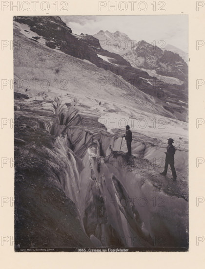 Crevasse am Eigergletscher, Two men at a crevasse in the Eiger Glacier, photograph, This photo is part of a cover., inscription: 'Gebr. Wehrli, Kilchberg, Zürich. 3665. Crevasse am Eigergletscher', Eiger, publisher: Gebrüder Wehrli, 1897 - 1920, photographer, Alps, Bernese Alps, paper, pasteboard, baryta paper, daylight collodion silver print, height 220 mm x width 163 mm, ravine, chasm, abyss, canyon, glacier, mountain climbing, mountaineering, A black and white photograph of a mountainous icy landscape showing a large glacier with deep crevasses and smooth, striated ice surfaces extending up a steep slope toward rocky, snow-dusted peaks in the background; two men in dark clothing and hats stand on the glacier near the edge of a deep fissure, one holding a long pole and the other holding a rope, both facing the crevasse; jagged rock faces and patches of snow appear on the higher slopes behind them, and the sky above is pale with scattered clouds.