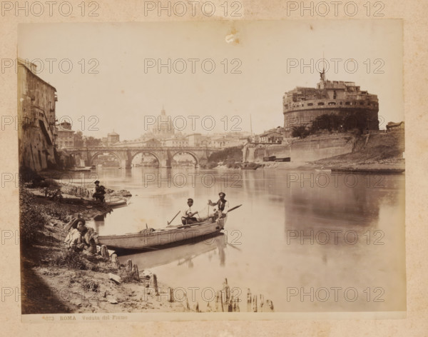 View of the Tiber River in Rome, album page, photograph, This photo is part of an album., inscription: '573 ROMA Veduta del Fiume', Rome, 1870 - c. 1895, photographer, paper, pasteboard, albumen print, height 200 mm x width 258 mm, prospect of city, town panorama, silhouette of city, canals, waters (in city), Tiber, A riverside scene shows calm water reflecting a stone bridge with multiple arches and buildings lining the far bank, including a large cylindrical fortress with battlements and a statue on its roof; nearer the left bank there are old multi-story buildings with shuttered windows and a sloped embankment leading to the water where small boats are moored and wooden stakes protrude from the muddy shore; several men sit and stand in and beside the boats, some holding oars or fishing nets, wearing hats and simple clothing; distant domed architecture rises behind the bridge, and trees and low walls dot the riverbanks under a pale sky.