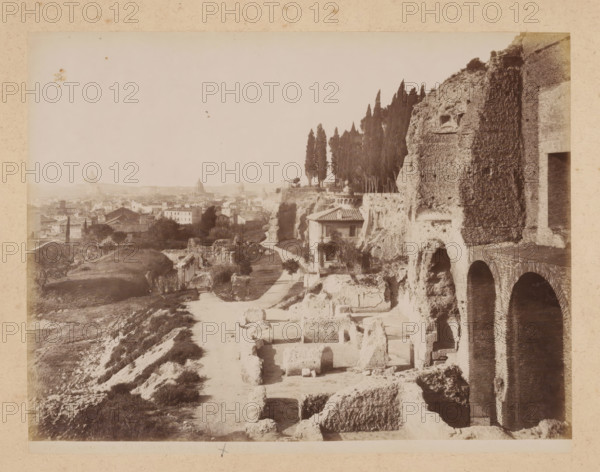 View of Rome from the Palatine Hill, album page, photograph, This photo is part of an album., inscription: '37. VEDUTA DI ROMA DAL PALATINO', Rome, 1870 - c. 1895, photographer, paper, pasteboard, albumen print, height 200 mm x width 258 mm, prospect of city, town panorama, silhouette of city, A sepia-toned view of an ancient stone ruin on a hillside with large arched openings and weathered brickwork along the right edge, terraced stone blocks and carved niches visible in the foreground, a dirt path winding past scattered rectangular stone remains toward a small building with a tiled roof set into the cliff, a line of tall, narrow cypress trees rising behind the cliff edge, cultivated plots and trees on lower slopes, a distant urban skyline of low buildings and domed rooftops across the horizon, and a pale sky filling the upper portion of the image.