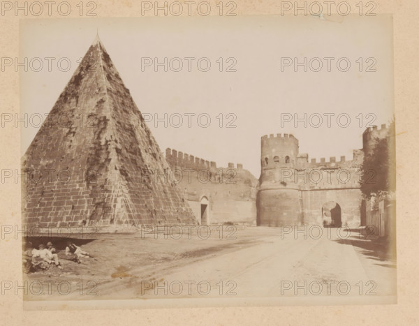 View of the Pyramid of Cestius and the Porta San Paolo in Rome, album page, photograph, This photo is part of an album., inscription: '137 PIRAMIDE DI GAIO CESTIO CON PORTA S. PAOLO', Rome, 1870 - c. 1895, photographer, paper, pasteboard, albumen print, height 201 mm x width 259 mm, city-gate, pyramid (marking grave), Porta San Paolo, Pyramid of Cestius, A large stone pyramid with weathered, patterned blocks fills the left side, showing a pointed top and dark streaks on its surface, while to the right a fortified stone wall with crenellations stretches toward cylindrical towers with rounded battlements and arched openings, one tower featuring small window-like apertures and another forming an arched gateway leading into a shadowed passage; a dirt road runs in front of the structures with faint wheel or track marks, a shadowed tree or foliage appears near the rightmost tower, and a group of people recline on the ground in the shaded area at the pyramid’s base.