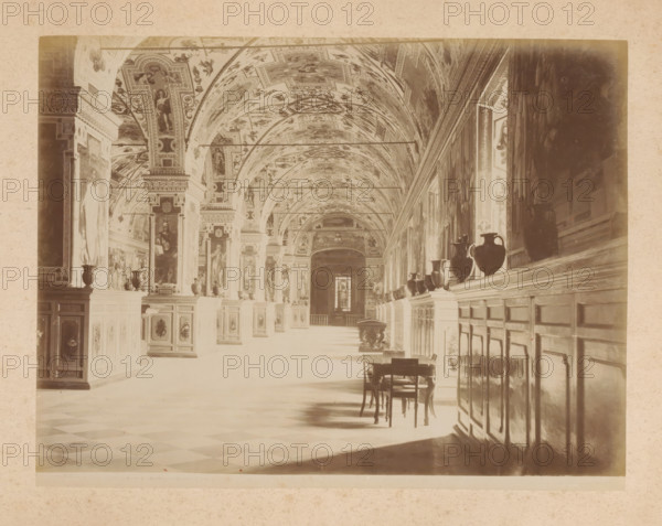 View of the Sistine Chapel in the Apostolic Palace in Vatican City, album page, photograph, This photo is part of an album., inscription: '385 SALA DELLA ... VATICANO', Sistine Chapel (Vatican), 1870 - c. 1895, photographer, Vatican City, Apostolic Palace, paper, pasteboard, albumen print, height 200 mm x width 260 mm, parts of church interior: chapel, A long ornate interior with a vaulted ceiling covered in painted panels and decorative motifs, tall pillars and arches lining a central corridor, framed paintings and sculpted figures set into wall panels, a row of dark vases displayed on a continuous ledge along one side, wooden cabinets or paneling beneath the ledge, a small wooden table and chairs placed on the tiled floor, checkerboard floor pattern visible in the foreground, soft light casting shadows across the floor and furnishings, and a distant doorway or window at the far end of the corridor.