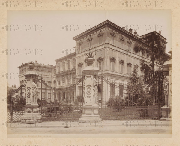 View of the Palazzo Barberini in Rome, album page, photograph, This photo is part of an album., inscription: '60 IL CAMPIDOGLIO', Rome, 1870 - c. 1895, photographer, paper, pasteboard, albumen print, height 199 mm x width 258 mm, palace, Palazzo Barberini, A sepia-toned photograph showing the façade of a large ornate building behind an iron fence, with a central corner section featuring shuttered windows and decorative window pediments, fluted pilasters, and a pronounced cornice; in front of the fence are two tall stone pillars topped with decorative urns and sculptural reliefs, and a curved wrought-iron gate with intricate scrollwork; behind the fence are planted shrubs and taller trees partially obscuring the right side of the building, and the street and sidewalk run across the foreground.