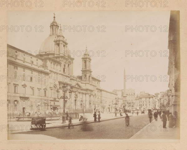 View of the Piazza Navona in Rome, album page, photograph, This photo is part of an album., inscription: '423 PIAZZA NAVONA', Rome, 1870 - c. 1895, photographer, paper, pasteboard, albumen print, height 200 mm x width 259 mm, square, circus, Piazza Navona, A wide sepia photograph showing a large ornate stone church with a prominent dome and flanking towers, detailed facades, and decorative cornices, a broad cobblestone street in front, ornate street lamps and a fenced plaza area with sculptures and fountains, groups of people standing and walking along the sidewalk and near the plaza, a cart on the street, rows of multi-story buildings receding into the distance, and an obelisk visible in the background under a pale sky.