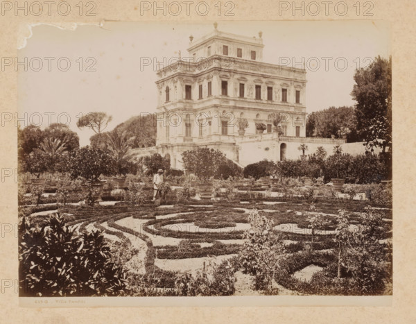 View of the Villa Doria Pamphilj in Rome, album page, photograph, This photo is part of an album., inscription: '615 G - Villa Pamfili', Rome, 1870 - c. 1895, photographer, paper, pasteboard, albumen print, height 198 mm x width 259 mm, villa, park, Janiculum, Villa Doria Pamphili, A sepia-toned photograph shows a formal garden with low clipped hedges arranged in concentric and curving patterns in the foreground, interspersed with gravel pathways and potted plants on raised bases. Behind the patterned beds, taller ornamental shrubs and small trees form a dense boundary, with a person wearing light clothing and a hat standing among the plants near a central path. A multi-story classical villa with ornate façade details, rows of windows, sculptural reliefs, decorative cornices, and a terrace with balustrade rises behind the garden. Mature trees, including a tall umbrella-shaped tree, frame the villa on either side, and potted plants sit on the villa's terrace and stair landing. The image...
