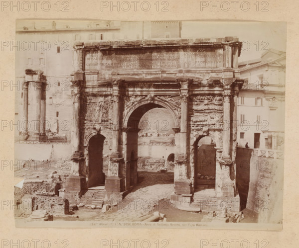 View of the Arch of Septimius Severus at the Roman Forum in Rome, album page, photograph, This photo is part of an album., inscription: '(Ed.ne Alinari) Pa Ia No 5834. ROMA - Arco di Settimio Severo, nel Foro Romano.', photographer: Fratelli Alinari (possibly), Rome, 1870 - c. 1895, paper, pasteboard, albumen print, height 192 mm x width 247 mm, triumphal arch, Arch of Septimius Severus, A large stone triumphal arch with three arched openings fills the foreground, showing worn columns, carved reliefs, and decorative moldings; the central arch is the tallest and flanked by paired columns with sculpted details, while the lower sections reveal stepped bases and eroded stonework. Behind and beside the arch are multi story buildings with plain facades and shuttered windows, and a tall isolated pair of standing columns is visible to the left. The ground in front of and beneath the arch consists of uneven cobbles, scattered stone fragments, and exposed foundations. A low railing runs along th...
