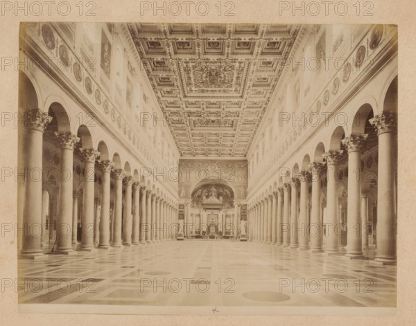 Interior of St. Paul Outside the Walls in Rome, album page, photograph, This photo is part of an album., inscription: '... FIORE LE MURA', St. Paul Outside the Walls, 1870 - c. 1895, photographer, Rome, Saint Paul Outside the Walls, paper, pasteboard, albumen print, height 199 mm x width 258 mm, interior of church, A wide interior view of a long, columned hall with two parallel rows of tall stone columns topped by ornate capitals and connected by rounded arches, a patterned floor with geometric shapes, a richly decorated coffered ceiling with square panels and an elaborate central design, circular medallion-like decorations along the upper walls, large arched openings above the columns, sculpted statues placed near the far end, and a decorated apse area with painted scenes and architectural ornamentation.