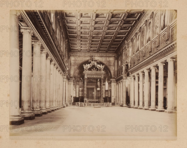 Interior of the Santa Maria Maggiore in Rome, album page, photograph, This photo is part of an album., inscription: '129 S. MARIA MAGGIORE', Rome, 1870 - c. 1895, photographer, paper, pasteboard, albumen print, height 202 mm x width 260 mm, interior of church, Santa Maria Maggiore (Rome), A wide interior view of a large hall with parallel rows of tall stone columns topped by ornate capitals on both sides leading toward a raised ornate altar area; a coffered decorative ceiling with square panels spans the space above, and high walls lined with framed arched panels and decorative molding run along the length; the altar area features sculptural figures, a canopy structure with columns, and religious furnishings, while the floor is open and patterned with rectangular stone sections and a few benches placed near the columns.