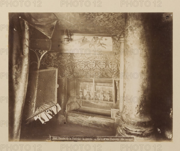 Grotte de la Nativité: la crêche, Cave of the Nativity: the manger, Cradle of Christ in the Church of the Nativity in Bethlehem, album page, photograph, This photo is part of an album., number: '893', photographer: Félix Bonfils, Bethlehem (Israel), 1867 - 1885, Palestine, paper, albumen print, height 218 mm x width 277 mm, interior of church, cradle, crib, Church of the Nativity, A dim, sepia-toned interior scene showing patterned fabric draped across the ceiling and walls, a hanging lamp suspended by chains, ornate carved columns with worn surfaces, a mesh-covered case or screen with decorative edging, a cushioned or padded surface beneath the mesh, and layered textiles with floral or damask motifs filling much of the space.