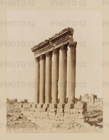 View of the pillars of the Temple of Jupiter in Baalbek, Lebanon, Balbek-Colonnes du temple du Soleil, album page, photograph, This photo is part of an album., number: '451', photographer: Félix Bonfils, Baalbek, 1867 - 1885, paper, albumen print, height 276 mm x width 216 mm, landscape with ruins, Temple of Jupiter (Baalbek), A row of tall stone columns with ornate capitals supports a partially preserved horizontal entablature, standing on a stepped stone platform amid scattered blocks and rubble, with a pale sky in the background and a lone small figure seated near the base of the structure.