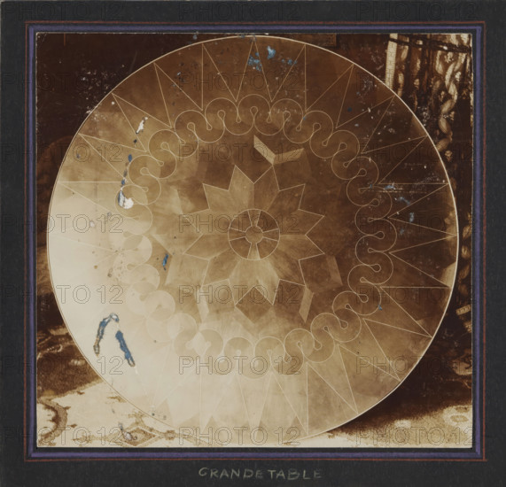 Tabletop designed by Mathieu Lauweriks, photograph, annotation: 'JM Lauweriks-, Ma...ts, 18, Düsseldorf, top of the large table', annotation: 'GRANDE TABLE', inscription: 'salon furniture executed in Cuban mahogany wood with gilded and chased brasswork, for Mr. JPW Schuurman, The Hague., Designed by JLM Lauweriks, architect., Executed by the firm Wed. C. Nederkoorn, furniture maker in Haarlem. + Sculpture, by MJ Hack in Amsterdam. + Brasswork by, F. Zwollo, Amsterdam +++++++++', commissioner (person who orders works): Mathieu Lauweriks, c. 1910, photographer, commissioner (person who orders works), paper, pasteboard, daylight collodion silver print, height 179 mm x width 187 mm, table, A close-up view of a circular tabletop with a decorative geometric pattern etched into its surface, showing radial star shapes and a scalloped inner ring with teardrop motifs, set against a dark rectangular frame with a thin purple inner border; the tabletop surface appears worn with patches of discolora...
