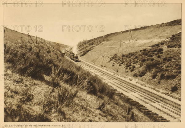 State Railways train on the tracks in the hills of Gelderland, The SS between the Gelderland hills, album page, photomechanical print, This print is part of an album., Gelderland, cliché maker: Nederlandsche Rotogravure Maatschappij, Leiden, in or before 1915, photographer, paper, rotogravure, height 161 mm x width 239 mm, railway, train, low hill country, A sepia-toned landscape showing railroad tracks running through a valley between low hills covered in grasses and shrubs, with a steam locomotive pulling cars and emitting a plume of smoke as it travels along the rails, wooden ties visible alongside the tracks, scattered telegraph poles standing on the hillsides, and dense vegetation in the foreground sloping down toward the tracks.