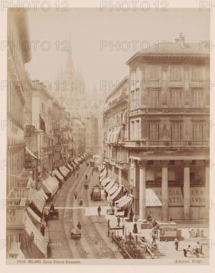 View of the Corso Vittorio Emanuele in Milan, Italy, Corso Vittorio Emanuele, Milano, photograph, number: '3830.', photographer: Giacomo Brogi, Milan, publisher: Edizioni Brogi, Florence, 1864 - 1881, Tuscany, Lombardy, pasteboard, paper, albumen print, height 330 mm x width 243 mm, urban housing, street, Corso Vittorio Emanuele II, A sepia-toned street scene showing a wide avenue flanked by tall multi-story buildings with shuttered windows and awnings, a large corner building with columns and a tiled roof on the right, tram tracks running down the center of the street, small horse-drawn carts and early motor vehicles and bicycles moving along the roadway, pedestrians walking on sidewalks and standing near storefronts, awnings and signs above ground-floor shops, scaffolding and construction activity with workers and materials visible near the bottom right, and a distant ornate cathedral façade rising above the street between the buildings.