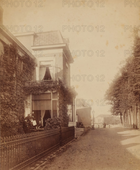 Street scene, with man, woman, and two children in front of a house (former title), Johannes Hendrikus Graadt van Roggen and his family in the doorway of their house at Lage Markt 33 in Nijmegen, photograph, Johannes Hendrikus Graadt van Roggen and his family in the doorway of their home at Lage Markt 33 in Nijmegen, annotation: 'Lange ?', stamp: 'W.Jvens (sic!), Photo aaf (?), Nijmegen', photographer: Wilhelm Ivens, Nijmegen, c. 1890 - c. 1915, paper, pasteboard, gelatin silver print, gelatin silver print on photographic paper, height 270 mm, width 220 mm, Person, Johannes Hendrikus Graadt van Roggen, A sepia-toned street scene showing the facade of a multi-story brick house partially covered in climbing vines, with tall windows and a small balcony with decorative railing and potted plants; curtains hang in the open windows and a group of people sit and stand on the ground-level porch area near a wrought-iron fence; a tree-lined street runs alongside the house with a lamp post and dis...