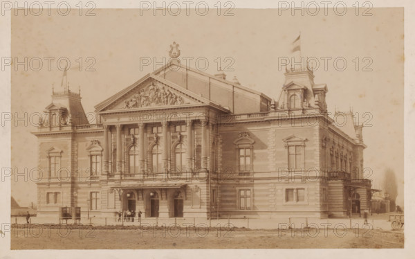 Concertgebouw, Amsterdam, photograph, viewed from the front right, the entire building is visible in the photo. Several figures are posing in front of the building., number: '75.', annotation: '160....illegible word', 1888-04-11 - c. 1915, photographer, paper, gelatin silver print, gelatin silver print on photographic paper, height 147 mm, width 240 mm x width 275 mm, height 172 mm, A large ornate stone building with a central pediment featuring a sculpted relief above a row of tall columns and arched windows, flanked by symmetrical towers with mansard roofs and decorative railings, two flags flying atop the roofline, a covered entrance with multiple doorways at ground level, small groups of people standing near the entrance, lamp posts and a low fence along the front, and a dirt area in the foreground.