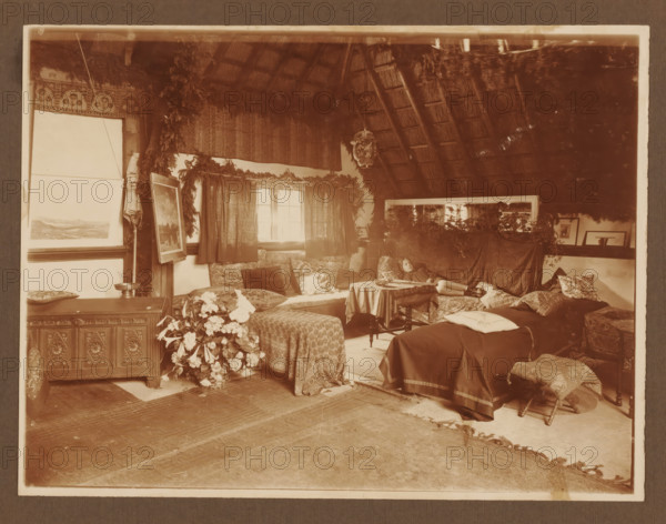 Girl lying on divan, bed in interior, ca. 1916, album page, photograph, ca. 1916. Interior with high thatched roof. Multiple beds, sofas, flowers and art on the walls and on the floor., c. 1916, photographer, paper, pasteboard, gelatin silver print, developing gelatin silver print on photo paper, height 180 mm x width 237 mm, height 220 mm x width 299 mm, A sepia-toned interior scene showing a cozy room with a thatched angled ceiling and exposed rafters, fabric draped and foliage hung along the walls and window area, a window with dark curtains and cushions on a cushioned bench beneath it, patterned cloths covering tables and seating, a small round table with a cloth and items on it, framed artworks leaning against the walls and resting on surfaces, a wooden cabinet with carved panels, a bouquet of flowers on a low table, assorted pillows and cushions scattered on benches and beds, a footstool with patterned upholstery, a large mirror behind foliage reflecting part of the room, and a p...