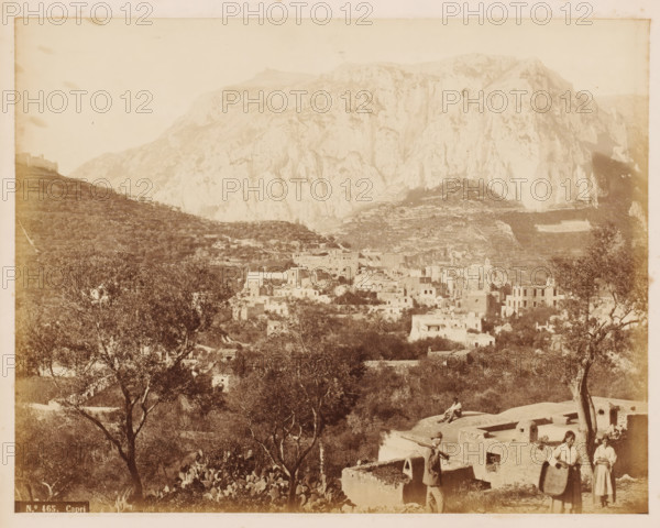 Capri, View of the city of Capri, photograph, number: 'No. 465', photographer: attributed to Roberto Rive, Isola di, 1860 - 1889, Attributed to, pasteboard, paper, albumen print, height 194 mm x width 244 mm, height 239 mm x width 277 mm, city-view in general, 'veduta', mountains, A sepia photograph of a hillside village with clustered stone and stucco buildings nestled among terraces and vegetation in front of a large rocky mountain; trees and low shrubs fill the foreground, with a few scattered cacti and a couple of figures near ruined low walls, one person standing and carrying a long pole and two women holding shallow baskets, and another figure seated on a flat roof; the mountains rise steeply behind the village with layered rock faces and light shadowing across the slopes, and a distant slope with terraced fields visible to the left.