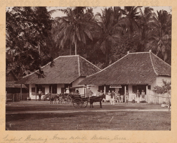 English boarding houses outside Batavia (present-day Jakarta), Java, Dutch East Indies, photograph, Dutch East Indies. Above the entrance of one of the houses hangs the sign: 'WJ Schinkel Boarding House'. In front of the houses, a couple, wagons, and people are posing. Palm trees in the background. Possibly an album page., inscription: 'English Boarding Houses outside Batavia Jarva. sic', c. 1900 - c. 1915, photographer, pasteboard, paper, gelatin silver print, height 161 mm, width 216 mm x width 289 mm, height 244 mm, West Java, Batavia, A sepia photograph shows a row of single-story buildings with tiled pitched roofs set against a backdrop of tall palm trees and dense foliage; in front of the buildings a group of people stand and sit, some wearing light-colored clothing and hats, others in darker garments, and several appear to be posed near or beside horse-drawn carriages with large wooden wheels; a wooden fence runs along one side of the scene and a dirt ground extends in the foreg...
