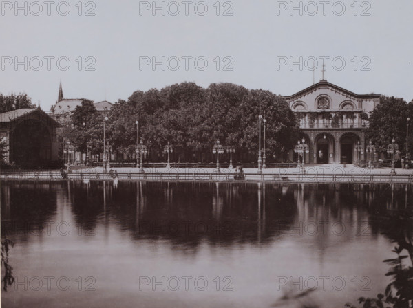 Kurgarten, Wiesbaden, Germany, photograph, Germany., inscription: '3. Wiesbaden, Kurgarten.', c. 1880 - c. 1900, photographer, pasteboard, paper, collotype, photoprint, height 205 mm, width 265 mm x width 330 mm, height 508 mm, Hessen, A wide view of a calm body of water reflecting a tree-lined promenade and ornate buildings, with dense trees centered behind a row of decorative lampposts and benches, flanked by a large arched open pavilion on the left and a symmetrical building with arched windows and balcony details on the right, all seen under a pale sky and mirrored in the smooth surface of the water.