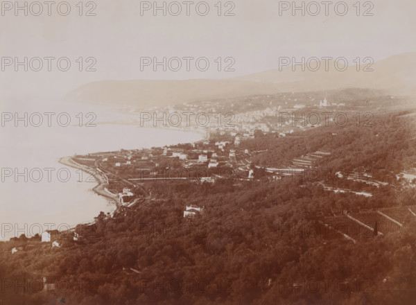 View from the Col de Poggio over the bay of San Remo, Italy, photograph, San Remo (Italian: Sanremo) is a seaside resort with approximately 50, 000 inhabitants in the Italian province of Imperia, Liguria region. The official name of the city is Sanremo. The spelling San Remo is often used, due to its resemblance to cities named after a saint., inscription: 'GJ phot.', inscription: '1963 SAN-REMO. - HOTEL ROYAL', photographer: Jean Gilletta, c. 1880 - c. 1900, paper, pasteboard, albumen print, height 213 mm, width 275 mm x width 330 mm, height 510 mm, San Remo, A sepia-toned aerial view shows a coastal town nestled between a shoreline and a series of hills, with a curved bay and a narrow strip of beach along the water's edge. Residential buildings and clustered houses sit near the shoreline and extend inland along winding roads and terraces, interspersed with patches of trees and cultivated plots. A dense expanse of wooded hillside occupies the foreground and lower slopes, with scattere...