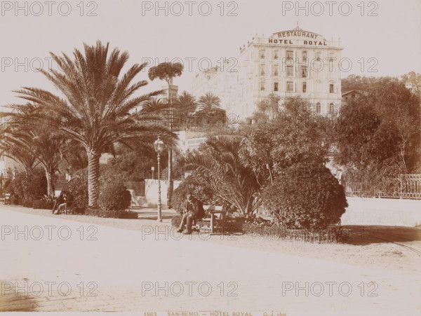 Hotel Royal, San Remo, Italy, photograph, San Remo (Italian: Sanremo) is a seaside resort with approximately 50, 000 inhabitants in the Italian province of Imperia, Liguria region. The official name of the city is Sanremo. The spelling San Remo is often used, due to its resemblance to cities named after a saint., inscription: 'GJ phot.', inscription: '2125 SAN-REMO - VUE PRISE DU COL DE POGGIO', photographer: Jean Gilletta, c. 1880 - c. 1900, paper, pasteboard, albumen print, height 213 mm x width 275 mm, height 510 mm x width 330 mm, A wide promenade lined with tall palm trees and trimmed shrubs runs in front of a multi-story classical building with many windows and balconies; a lamppost stands near a bench where a person wearing a coat and hat sits, and another person sits on a bench further left partially shaded by palm fronds; dense foliage and bushes fill the foreground and frame the building, and a decorative metal fence is visible at the right edge.