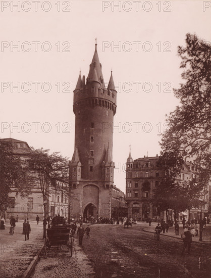 Eschenheimner Turm, Frankfurt-am-Main, Germany, photograph, Germany., caption: 'Frankfurt', c. 1875 - c. 1900, photographer, paper, pasteboard, collotype, photoprint on photo paper on cardboard, height 274 mm, width 208 mm x width 490 mm, height 330 mm, Hessen, Frankfurt am Main, A tall cylindrical tower with conical turrets and a large arched gateway stands at the center of a street scene, flanked by multi-story buildings with ornate facades and a cluster of trees with leafy branches on the right; cobblestone streets fill the foreground with tram tracks running down the middle, horse-drawn carriages parked near the curb, and numerous people walking or standing in period clothing scattered across the scene.