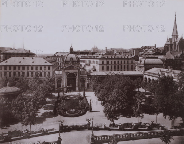 Kochbrunnen, Wiesbaden, Germany, photograph, View from above of a park and a Neoclassical building in the background, a church on the right., inscription: '4. Kochbrunnen-Anlagen.', c. 1890 - c. 1900, photographer, pasteboard, paper, collotype, photogravure, height 212 mm, width 267 mm x width 330 mm, height 510 mm, Hessen, A sepia-toned cityscape viewed from above shows a formal garden in the foreground with symmetrical flowerbeds, trimmed hedges, and pathways flanked by trees and lampposts, leading to a stone entrance terrace with balustrades and decorative urns; beyond the garden stands an ornate building with a large central archway, a domed roof topped by a small cupola and sculptural figures, flanked by adjacent buildings with rows of windows and mansard roofs; to the right a building with a curved roof and sculpted pediment sits near a larger church with a tall pointed spire and buttressed walls, while a dense cluster of rooftops, chimneys, and distant towers fill the midground ...