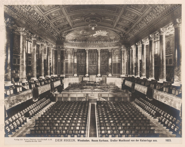 Der Rhein. Neues Kurhaus, Grosser Musiksaal von der Kaiserloge aus, Wiesbaden, Germany, photograph, Germany., caption: 'DER RHEIN. Wiesbaden. Neues Kurhaus. Grosser Musiksaal von der Kaiserloge aus.', name: 'Neue Photographische Gesellschaft AG, Steglitz-Berlin 1907', number: '1623.', photographer: Neue Photographische Gesellschaft, Berlin, 1907, pasteboard, paper, albumen print, albumendruk, height 192 mm, width 239 mm x width 328 mm, height 510 mm, Hesse, A large ornate concert hall with rows of seating arranged facing a raised stage area, flanked by tall decorative columns with carved capitals and ornate paneling along the walls, a curved apse behind the stage featuring intricate relief panels and decorative doors, an elaborately coffered ceiling with sculpted moldings and a central chandelier, balcony seating with carved balustrades on both sides, and many wooden chairs arranged on the stage and in the auditorium.