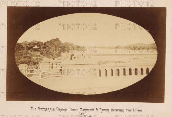 Fitgerald Bridge, Bund Gardens and Mula Mutha river, Pune, Maharashtra, India, photograph, Oval photo of a bridge with river and a park on the left. Poona, also called Pune and Puna is an Indian city not far from Mumbai., inscription: 'The Fitzgerald Bridge + Bund Gardens + River shweing the Bund, Poona', inscription: 'THE FITZGERALD BRIDGE, BUND GARDENS & RIVER SHEWING THE BUND., Poona.', photographer: X photo., c. 1875 - c. 1900, An oval sepia photograph framed by a dark rectangular border shows a riverside scene with a low balustrade in the foreground featuring decorative cutouts and carved circular motifs, a set of railings and a small stair or platform area, a row of trees and dense foliage to the left with a pavilion topped by a pointed roof visible among the trees, a low embankment leading to a wide calm river that stretches toward distant low hills under a pale sky, and faint signs of wear and spotting on the photographic surface.