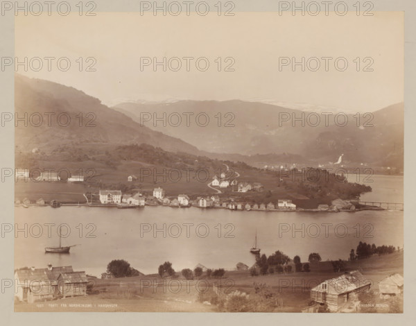 Parti fra Nordheimsund, Hardangerfjord ?, Norway, photograph, Norway., inscription: 'K. KNUDSEN, BERGEN', caption: '1027. PARTI FRA NORDHEIMSUND I HARDANGER.', photographer: Knud Knudsen, c. 1880 - c. 1900, paper, pasteboard, albumen print, height 209 mm, width 272 mm x width 402 mm, height 316 mm, A panoramic view shows a calm body of water in the foreground with two sailboats anchored near the shore, a shoreline lined with small wooden boathouses and larger white buildings, gently rolling grassy land with scattered trees and fences rising behind them, a winding road leading up to more clustered houses spread across the meadow, steep wooded hills and terraced slopes rising further back, distant mountains with pale snow patches near their peaks, and a bridge connecting a small promontory to the far shore.