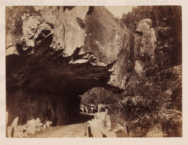 Parti ved halvtunnellen I Bratlandsdalen, Suldal, Norway, photograph, Tunnel of overhanging rocks., inscription: 'K. Knudsen, Bergen.', inscription: '8380. Parti ved halvtunnellen I Bratlandsdalen, Suldal', photographer: Knud Knudsen, Bergen, c. 1880 - c. 1900, paper, pasteboard, albumen print, height 160 mm, width 217 mm x width 310 mm, height 402 mm, A sepia-toned photograph showing a rocky cliff face with a dark overhanging ledge above a narrow dirt road, jagged rock formations lining the road and a low stone barrier along the roadside, a few standing figures on the road near the stone barrier, a steep wooded slope with trees and shrubs rising beside the cliff, and textured rock surfaces with light and shadow across the scene.