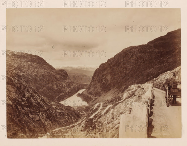View from Reldalsfjeldet with mountains around it, Norway, photograph, View over a deep valley surrounded by mountains, taken from a road with a cart next to it. Photographer appears to have dismounted., inscription: 'View from Reldalsfjeldet with mountains around it', inscription: 'K. Knudsen, Bergen.', photographer: Knud Knudsen, Bergen, c. 1880 - c. 1900, paper, pasteboard, albumen print, height 163 mm, width 218 mm x width 310 mm, height 386 mm, A sepia landscape photograph showing a narrow valley between steep rocky cliffs with a winding road carved along the cliffside and a river or lake running through the valley floor, a stone barrier lining the road in the foreground, a vehicle partially visible on the road at the right edge, and distant mountains and cloudy sky in the background.