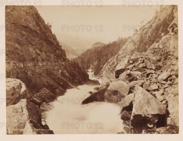 Parti Af Oddedalen, Norway, photograph, View of a wild flowing river between mountains and rocks, Norway., inscription: 'Udisgt fra Reldalsfjeldet nedover gaarsbottnen mod folgffonden', inscription: 'K. Knudsen, Bergen.', photographer: Knud Knudsen, Bergen, c. 1880 - c. 1900, paper, pasteboard, albumen print, albumendruk, height 163 mm, width 216 mm x width 316 mm, height 400 mm, A sepia-toned landscape showing a rocky river gorge with steep cliff faces on both sides and large boulders scattered along the riverbank, water flowing through the narrow channel appearing smooth and misty, coniferous trees clinging to the slopes and ridge lines, a rough stone retaining wall or path built into the cliff on the left side, distant hazy hills fading into the sky, and a foreground of angular rocks and ledges descending toward the water.