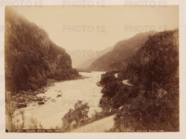 Ryfylke, Mellem-Osen, Sand, photograph, View of a wide river, Norway., caption: '2877. Ryfylke, ', inscription: 'Rich. Andvord. Eneret 1892.', publisher: Richard Andvord, photographer: attributed to Axel Lindahl, in or before 1892, Attributed to, paper, pasteboard, albumen print, albumendruk, height 165 mm, width 226 mm x width 310 mm, height 400 mm, Norway, A sepia-toned landscape photograph showing a river flowing between steep, forested cliffs, with rocks and boulders along the riverbanks and in the water, trees and dense vegetation covering the slopes on both sides, a winding dirt road hugging the right-hand cliff edge, misty or hazy distant hills receding into the background, and overall muted tones and soft contrast across the scene.