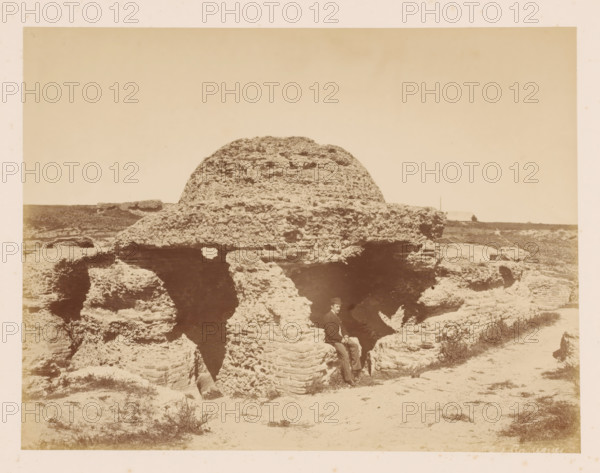 Canopy, house of wood stones?, with a man in front of it, photograph, with a man in front of it., inscription: 'Ph. J Carrianes ?', c. 1870 - c. 1900, photographer, pasteboard, paper, albumen print, height 197 mm, width 262 mm x width 443 mm, height 325 mm, A sepia photograph showing a large, rounded rock formation with layered, weathered textures and several deep crevices; the rock occupies the central foreground and middle distance, with smaller rocky outcrops and patches of sparse vegetation around its base and extending into the background; a man wearing a dark jacket, light trousers, and a hat is seated in a recess of the rock near the lower center of the image; the horizon and flat landscape stretch behind the rock under a clear, pale sky.