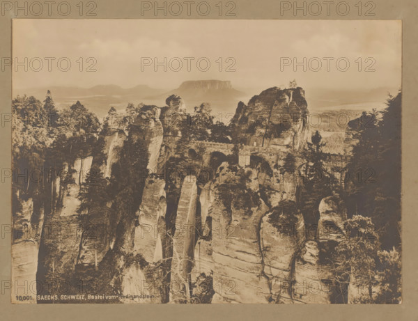 Bastei from the Ferdinandstein, Saxon Switzerland, Germany, photograph, Saxon Switzerland (Saxon Switzerland) is a former district (Landkreis) in the German state of Saxony and also the name for the region in the Elbe Sandstone Mountains between Dresden and the Czech border. Here we see the 'Bridge of Bastei', a natural wonder of several striking rock peaks with a man-made stone bridge., number: '1903', inscription: '10, 001 SAECHS. SCHWEIZ, Bastei vom Ferdnindstein.', check stamp: 'Th. ...-maier', 1903, photographer, pasteboard, paper, gelatin silver print, developing gelatin silver print, height 169 mm, width 230 mm x width 381 mm, height 320 mm, A sepia-toned photograph showing a rocky landscape with tall vertical sandstone pillars and cliffs, many topped with trees and vegetation, and a stone bridge spanning between rock formations; the foreground displays steep weathered rock faces with visible striations and sparse trees growing from crevices, while the midground features cluster...