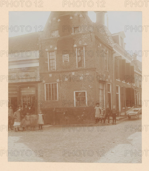 House in Dokkum, photograph, annotation: 'Dokkum House April 1911, (Friesland), Dokkum (Friesland)', (Friesland)', Dokkum, photographer: AJM Mulder (possibly), 1911, AJM Mulder, photographer, Attributed to, paper, pasteboard, daylight collodion silver print, height 171 mm x width 150 mm, urban housing, street (city(-scape) with figures, staffage), A faded sepia photograph showing a row of brick buildings with tiled roofs and multiple windows, some with shutters, and a decorative stone or plaster panel on one facade; people stand and blur in front of the buildings, including children and adults wearing period clothing and hats, with a small group clustered near a doorway and a few individuals posed at the corner; a wooden wheelbarrow rests on the cobbled street beside the buildings, and the street surface extends into the foreground.