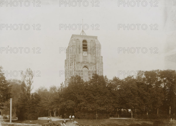 View of the Oldehove with nearby park in Leeuwarden, photograph, Leeuwarden, 1890 - 1910, photographer, paper, pasteboard, daylight collodion silver print, height 123 mm x width 170 mm, height 144 mm x width 194 mm, parts of church exterior and annexes: tower, A tall stone tower with arched openings and a pointed finial rises above a dense line of trees, the tower showing vertical brick patterns and a large arched recess near its base; in front of the trees a grassy area contains a few people standing and sitting along a low stone edge and a small rectangular water feature, while tall slender trees flank the scene and the sky above is pale and empty.
