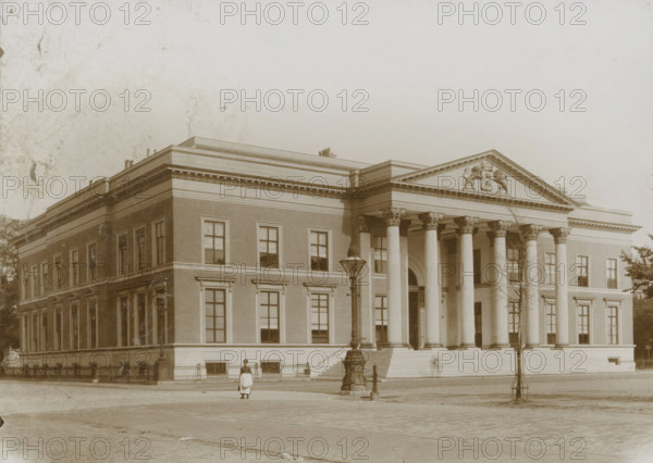 Palace of Justice in Leeuwarden, photograph, Leeuwarden, 1890 - 1910, photographer, paper, pasteboard, daylight collodion silver print, height 125 mm x width 173 mm, height 144 mm x width 194 mm, court building, A large classical stone building with a wide flight of steps leading to a portico supported by tall fluted columns is shown, the portico topped by a triangular pediment decorated with sculpted figures. The façade features rectangular windows set into brickwork with stone trim, a decorative cornice running along the roofline, and ornate capitals on the columns. A lamppost stands on the pavement near the steps, and a solitary person wearing a light-colored dress and apron is standing in front of the building. Sparse trees and a low fence are visible along the building's base, and the foreground shows a cobblestone street. The image has an overall sepia tone and some visible surface blemishes.