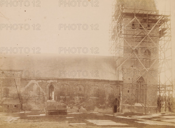 North side of the St. Vitus Church in Stiens, during restoration work, photograph, annotation: 'Ned. Herv Kerk te Stiens fr (Fr.), North side, Nov 1898', annotation: 'Leeuwarderadeel, Stiens', Stiens, photographer: AJM Mulder (possibly), 1898, Attributed to, paper, pasteboard, albumen print, height 164 mm x width 225 mm, church (exterior), Sint-Vituskerk (Stiens), A sepia-toned photograph showing a large brick building with arched windows and a steep roof, a tall tower at one end surrounded by wooden scaffolding, a small tiled-roof outbuilding near the corner, visible grave markers or flat stones on the grassy foreground, a wooden bench and some construction materials near the base of the building, and a faintly mottled sky above.