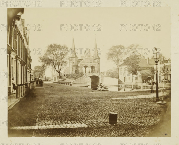 View of Sneek with the city side of the Sneeker Waterpoort, photograph, annotation: 'f1, - ..., Sneek', Sneek, 1870 - 1900, photographer, paper, pasteboard, albumen print, albumendruk, height 145 mm x width 182 mm, height 255 mm x width 286 mm, city-gate (city(-scape) with figures, staffage), Waterpoort (Sneek), A wide cobblestone street slopes toward a stone arch bridge flanked by two tall pointed towers, with trees on either side and people visible on the bridge and along the banks, a canal running beneath the bridge where a small boat is moored, brick row houses lining the left side of the street with several figures standing near doorways, a lamppost in the foreground on the right, low buildings and more trees beyond the bridge, and a pile of sacks near the canal edge.