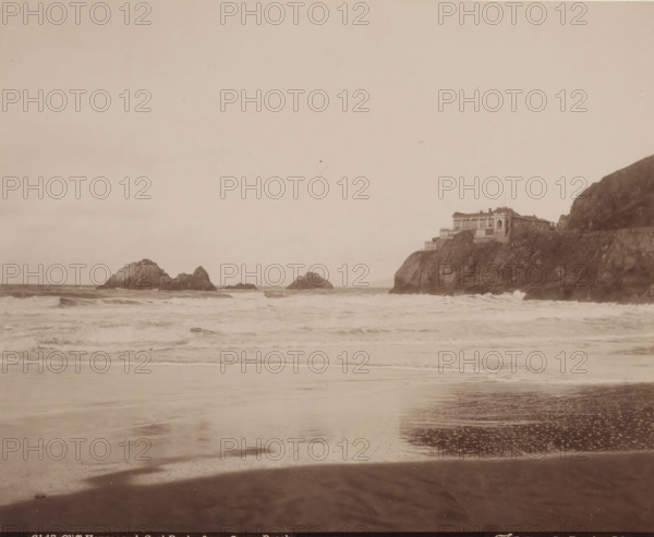 House on rock, Ocean Beach, California, United States, photograph, United States., inscription: '6147 Cliff House. and Seal Rocks from Ocean Beach, Faber Photo, San Francisco, Cal.', photographer: Isaiah West Taber, c. 1881 - c. 1910, pasteboard, paper, albumen print, albumendruk, height 199 mm x width 245 mm, height 275 mm x width 350 mm, North America, A wide seascape with waves rolling toward a sandy shore in the foreground, rocky outcroppings rising from the water toward the center, and a rocky cliff on the right topped by a multi-level building with terraces and railings; the sky above is pale and mostly clear.
