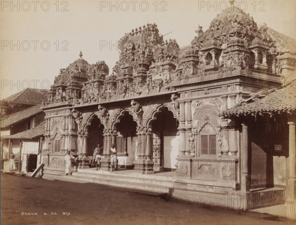 Hindu temple with priests, Colombo, Sri Lanka, photograph, Temple, Ceylon ? (current Sri Lanka)., name: 'Skeen & Co.', number: '873', photographer: WLH Skeen & Co., c. 1875 - c. 1910, paper, albumen print, height 213 mm x width 275 mm, South Asia, A stone temple facade with ornate carved arches and columns topped by sculpted figures and decorative domes occupies the foreground, showing layered reliefs of human and animal forms across the upper frieze and roofline; tiled sloping roofs of adjacent structures flank the temple on either side, and a wide stepped platform runs along the base of the facade; several people in traditional wrap garments stand and lean near the columns and steps, and shadows fall across the street in front of the temple.